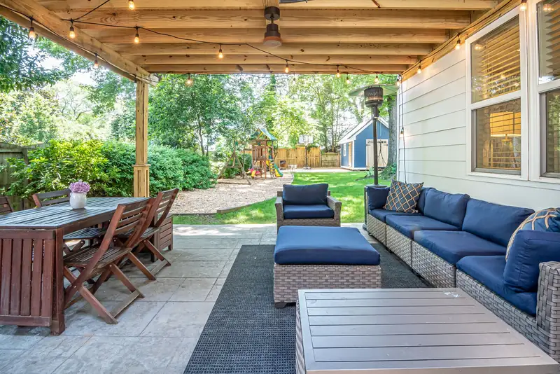 Covered patio with paved tile flooring, wooden dining table, blue sectional sofa, and a lawn with a play set behind