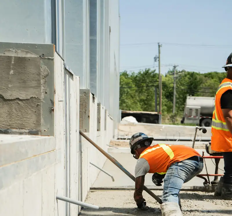 Construction worker in a high-visibility vest finishing a freshly poured concrete slab, face not shown