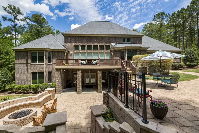 Paver patio behind a brick house with a stone fire pit, wooden chairs, umbrella table, and wicker furniture