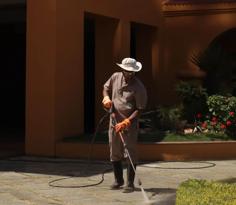 Worker in hat and gloves pressure-washing a paver patio in front of a stucco house, face shadowed and not the focus